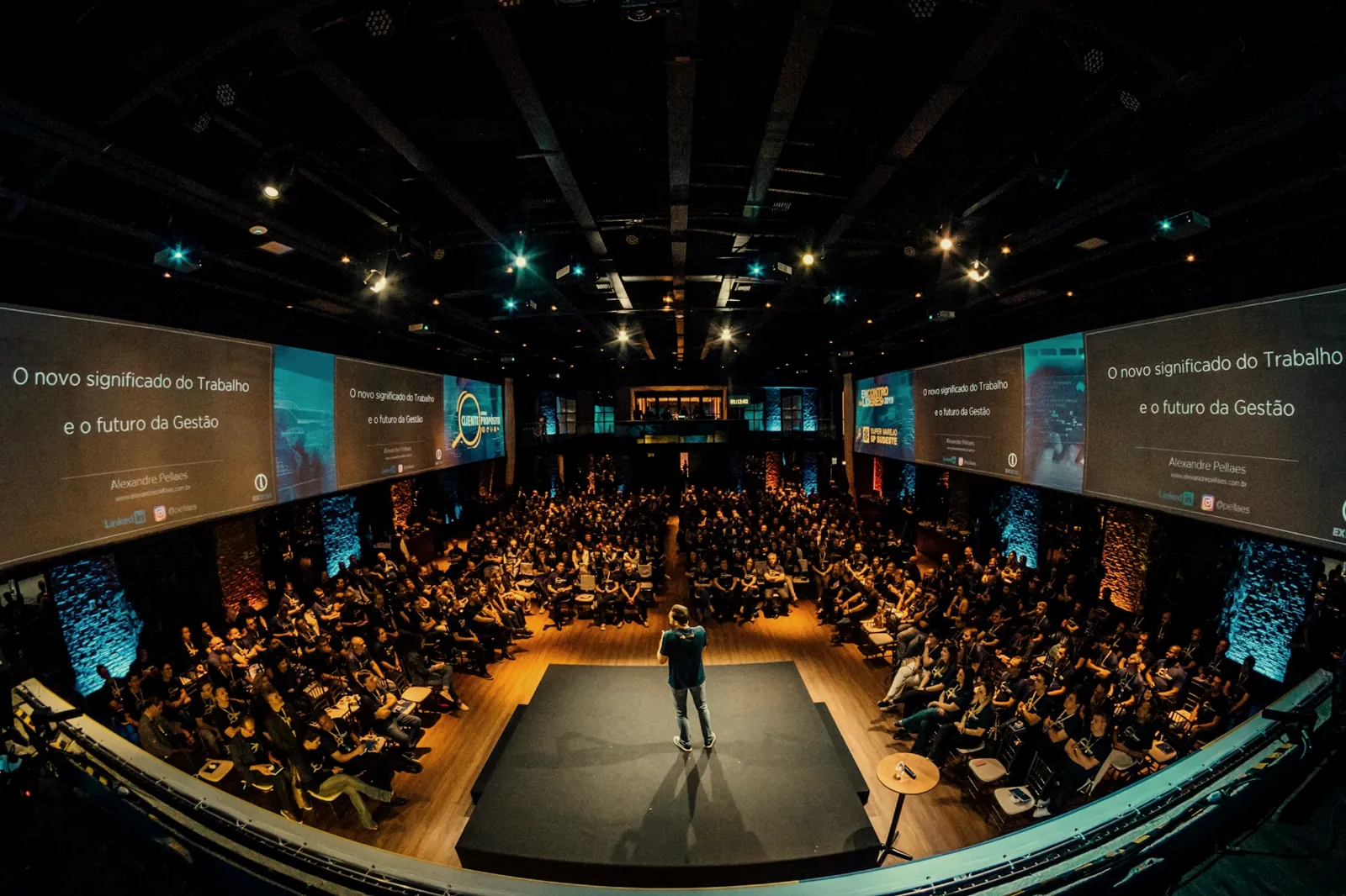 Palestrante no palco de um evento ao vivo, com telões e plateia cheia
